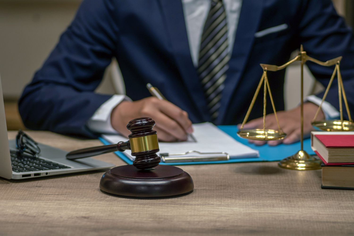 A lawyer writes notes at a desk with a gavel, scales of justice, books, and a laptop.